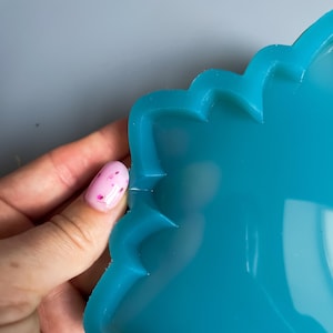 Pu&ograve; includere: Primo piano di uno stampo in resina color turchese con bordo smerlato. Lo stampo &egrave; tenuto da una mano con un'unghia rosa con un disegno floreale. Lo stampo &egrave; liscio e lucido.