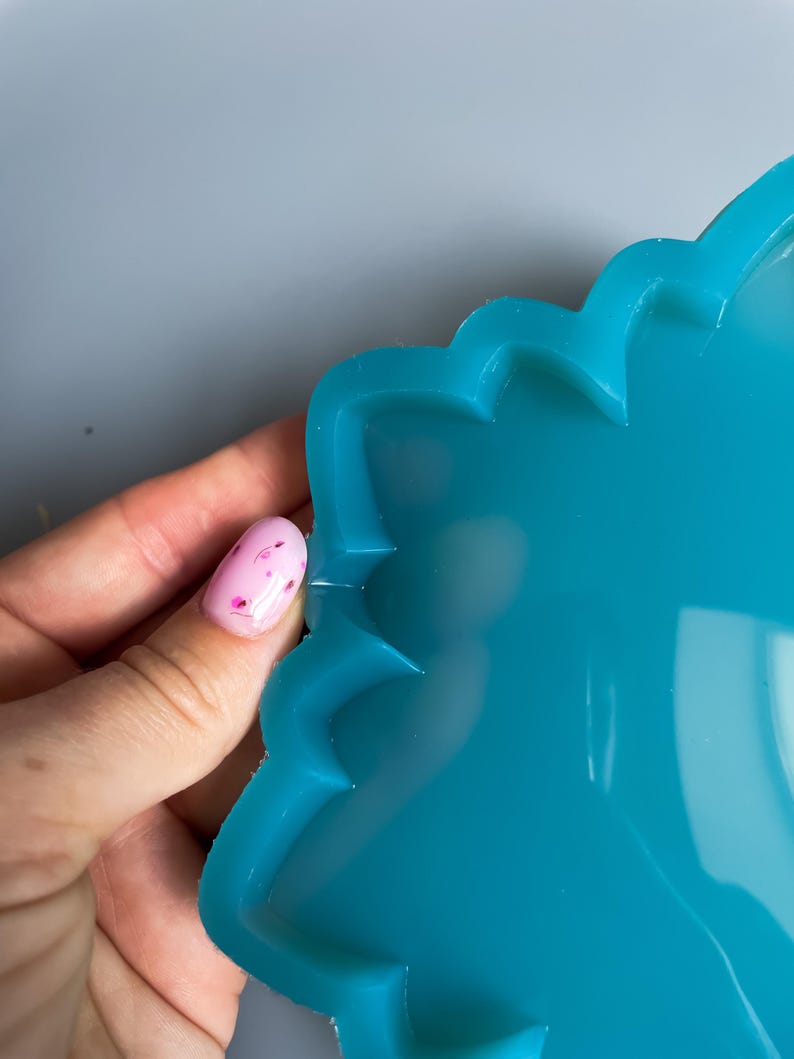 Pu&ograve; includere: Primo piano di uno stampo in resina color turchese con bordo smerlato. Lo stampo &egrave; tenuto da una mano con un'unghia rosa con un disegno floreale. Lo stampo &egrave; liscio e lucido.