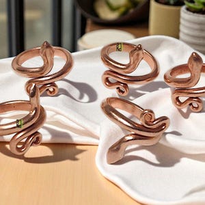 Puede incluir: Anillos de servilleta de metal color cobre con diseño de serpiente. Los anillos están enrollados con una cabeza de serpiente en la parte superior. Los anillos están sobre una tela blanca, con una superficie de madera clara en primer plano.