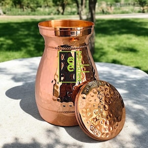 May include: A copper-coloured vase with a hammered texture and a matching lid. The vase has a wide base, a narrow neck, and a flared rim. The lid is round and has a hammered texture. The vase is sitting on a stone surface.