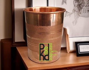 Könnte beinhalten: Ein kupferfarbener Metalleimer mit horizontalen geätzten Linien. Der Eimer hat einen glänzenden Rand und eine matte Oberfläche am Körper. Das Logo "PKD peekhdee" befindet sich auf der Vorderseite. Der Eimer steht auf einer Holzoberfläche.