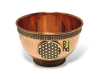 May include: A copper-colored bowl with a decorative Greek key pattern around the rim and base. A circular, gold-colored design is centered on the bowl's front. The bowl has a polished, reflective interior and a wide, open top.