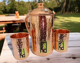 Puede incluir: Una jarra de metal martillado de color cobre con tapa, asa y dos vasos a juego. Cada pieza tiene un logotipo verde y negro. El juego se exhibe sobre una superficie de madera al aire libre, con una mesa de picnic y árboles al fondo.