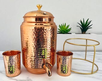 Può includere: Un set di erogatore d'acqua in rame con una texture martellata, composto da un grande contenitore con coperchio, due tazze abbinate e un supporto dorato. Il set include un rubinetto ed è esposto con piccole piante in vaso.