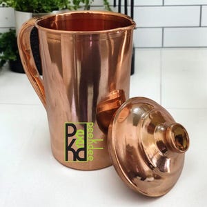 Puede incluir: Una jarra de cobre con tapa a juego. La jarra tiene un asa y una abertura ancha. La tapa es redonda con un pequeño pomo. La jarra y la tapa son de cobre brillante. La jarra tiene el texto "pkd" en un cuadrado verde.