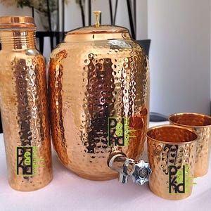 May include: A set of copper-colored drinkware, including a water bottle, a large hammered jug with a lid, two small cups, and a spigot. The items have a textured surface and a logo in green and black.