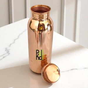 May include: A copper water bottle and lid. The bottle is cylindrical with a wider base and a narrow neck. The lid is round and sits beside the bottle. The bottle is made of shiny copper. The bottle's capacity is approximately 750 ml.
