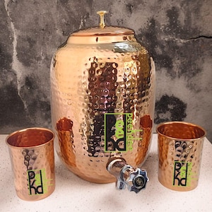 May include: A copper-colored water dispenser with a hammered texture, a lid, and a spigot. Two matching copper cups are on either side. The dispenser and cups have a logo in green and black. The background is a textured gray wall.