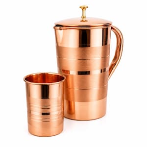 May include: A polished copper pitcher with a matching tumbler. The pitcher features a lid with a gold-toned knob and horizontal bands of brushed and shiny finishes. The cylindrical tumbler has similar horizontal bands around its exterior surface.