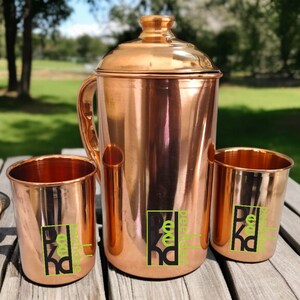 Puede incluir: Una jarra de agua de cobre con tapa, asa y dos vasos de cobre a juego. La jarra y los vasos tienen un acabado brillante y un logotipo en verde y negro. Los artículos están sobre una mesa de madera al aire libre.