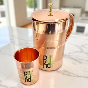 May include: A copper-coloured jug with a matching lid and a handle, next to a copper-coloured tumbler. The jug and tumbler have a logo printed on them. The jug is approximately 25 cm tall.