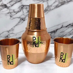 Könnte beinhalten: Ein kupferfarbenes Getränkeset mit gehämmertem Muster. Das Set umfasst einen hohen Krug mit Deckel und zwei passende Becher. Jedes Teil hat ein grünes und schwarzes Logo. Das Set ist zum Servieren von Getränken bestimmt.