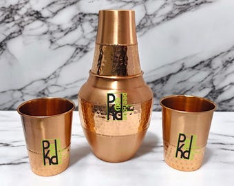 May include: A copper-colored drinkware set featuring a hammered texture. The set includes a tall, lidded pitcher and two matching cups. Each piece has a green and black logo. The set is designed for serving beverages.