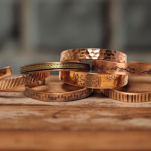 May include: Several copper bracelets with diverse designs, such as etched patterns, hammered textures, and mountain silhouettes. The bracelets are arranged on a wooden surface, highlighting their warm, metallic hues.