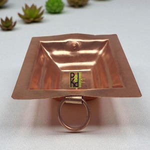Puede incluir: Un cuenco cuadrado de color cobre con un asa de anillo. El cuenco tiene un borde ligeramente elevado y un pequeño asa circular unida a la parte inferior. El cuenco está hecho de un material metálico brillante y tiene un diseño simple y elegante.