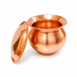 May include: A polished copper pot with a rounded body and a wide, flared rim, accompanied by a matching circular lid placed upright to the left. The metal surface features a subtle horizontal brushed texture and a reflective finish.
