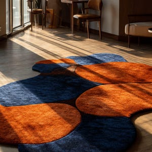 May include: A contemporary rug with an abstract design. The rug has navy blue and orange sections, forming a distinctive pattern. It's placed on a wooden floor, with sunlight coming in from a window. The rug's design is eye-catching.