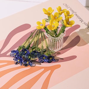 Könnte beinhalten: Eine kleine Glasvase enthält gelbe Glasblumen mit blauen Zentren. Mehrere blaue Glasblumen sind in der Nähe verstreut. Die Blumen befinden sich auf einem rosa-pfirsichfarbenen Hintergrund. Die Blumen sind ein dekorativer Gegenstand.