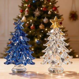 Op de afbeelding: Twee decoratieve glazen kerstbomen. Eén is diepblauw en de andere is helder met een gouden ster erop. Beide hebben een facetontwerp en staan op een heldere basis. De achtergrond toont een versierde kerstboom.