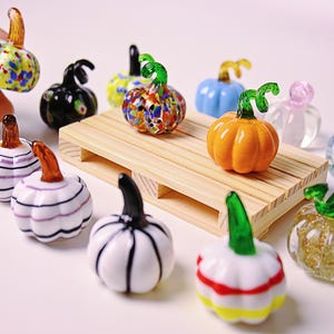 Könnte beinhalten: Eine Sammlung von Miniatur-Glas-Kürbissen in verschiedenen Farben und Designs. Einige Kürbisse sind orange, blau und weiß, mit grünen oder braunen Stielen. Ein Kürbis wird von einer Hand gehalten. Die Kürbisse sind auf einer kleinen Holzpalette angeordnet.