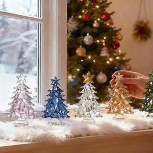 May include: A collection of decorative glass Christmas trees in various colors, including pink, blue, clear, gold, and green, each topped with a star. They are displayed on a windowsill with a snowy outdoor view.