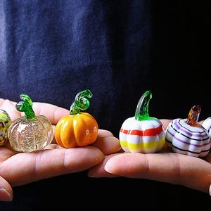 Pode incluir: Uma coleção de seis mini abóboras de vidro em várias cores e padrões. Cada abóbora tem um caule verde ou laranja. As abóboras são exibidas em mãos abertas contra um fundo escuro, mostrando seus detalhes e cores vibrantes.