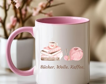 May include: A white ceramic mug with a pink interior and handle, featuring a graphic of a stack of books topped with a coffee cup, a ball of pink yarn, and a knitting needle, with the text "Bücher. Wolle. Kaffee." printed in brown lettering.