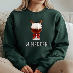 May include: A dark green crewneck sweatshirt featuring a graphic of a wine glass adorned with brown reindeer antlers, a string of small yellow fairy lights, and a red bow at the base. The text WINEDEER is printed in white below the wine glass graphic.