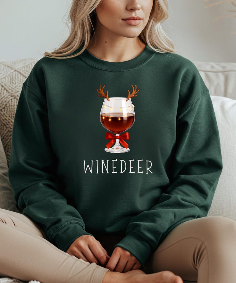 May include: A dark green crewneck sweatshirt featuring a graphic of a wine glass adorned with brown reindeer antlers, a string of small yellow fairy lights, and a red bow at the base. The text WINEDEER is printed in white below the wine glass graphic.