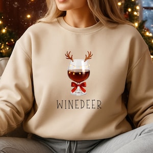 May include: A beige crewneck sweatshirt featuring a graphic of a wine glass with brown reindeer antlers, a string of small yellow lights, and a red bow. Below the graphic, the text WINEDEER is printed in a simple black sans-serif font.