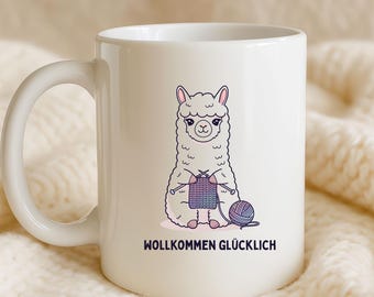May include: A white ceramic mug featuring a cartoon illustration of a fluffy white llama knitting with purple and blue yarn. Below the llama, the text WOLLKOMMEN GLÜCKLICH is printed in black capital letters. The mug has a curved handle.