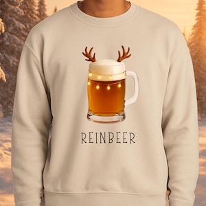 May include: A beige crewneck sweatshirt featuring a graphic of a glass beer mug with foam, brown reindeer antlers, and glowing string lights wrapped around the top. The text REINBEER is printed in black capital letters below the beer mug graphic.