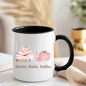 May include: A white ceramic mug with a black interior and handle, featuring a graphic of a stack of pink books topped with a coffee cup, a ball of pink yarn with a needle, and the text "B&uuml;cher. Wolle. Kaffee." printed in brown lettering.