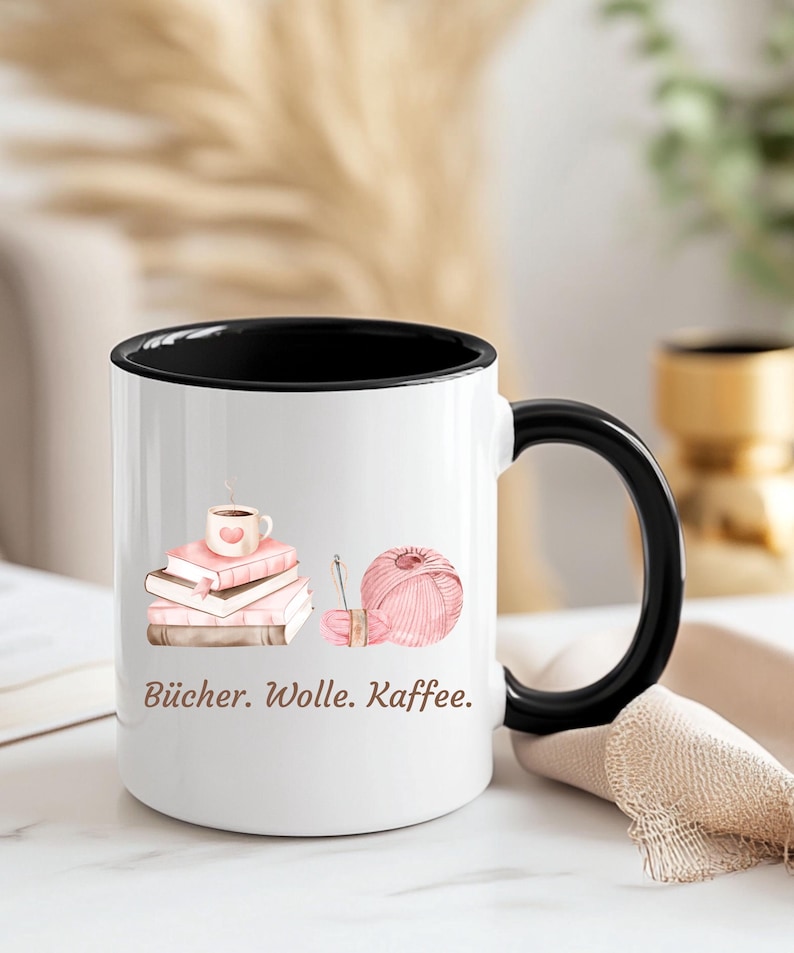 May include: A white ceramic mug with a black interior and handle, featuring a graphic of a stack of pink books topped with a coffee cup, a ball of pink yarn with a needle, and the text "B&uuml;cher. Wolle. Kaffee." printed in brown lettering.