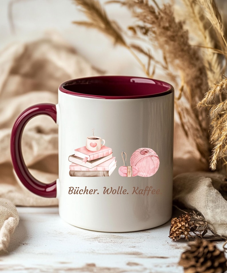 May include: A white ceramic mug with a deep burgundy interior and handle. The exterior features a graphic of stacked pink books topped with a small coffee cup, alongside a ball of pink yarn. The text "B&uuml;cher. Wolle. Kaffee." is printed below.