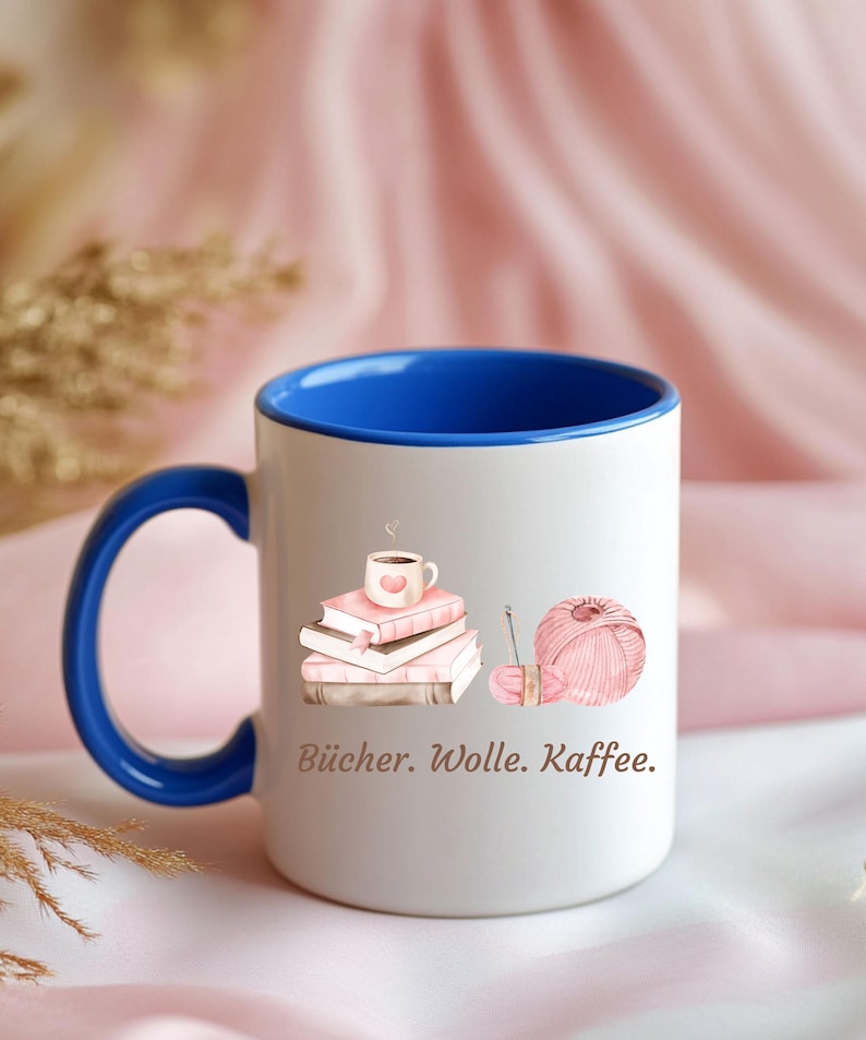 May include: A white ceramic mug with a vibrant blue interior and handle. The exterior features a graphic illustration of a stack of books topped with a coffee cup, alongside a ball of pink yarn with a needle. The text reads: B&uuml;cher. Wolle. Kaffee.