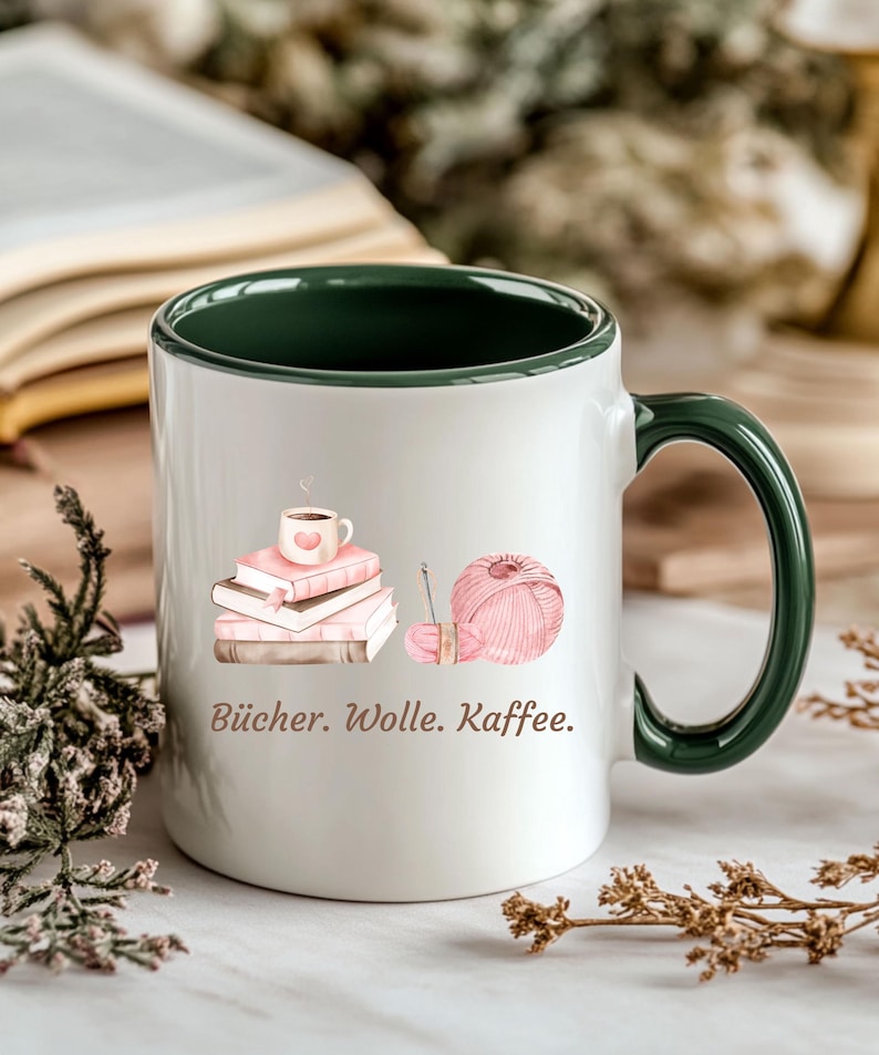 May include: A white ceramic mug with a dark green interior and handle. The exterior features a graphic of three stacked books topped with a coffee cup, a ball of pink yarn with a needle, and the text "B&uuml;cher. Wolle. Kaffee." printed in brown.