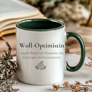 Può includere: Una tazza in ceramica bianca con interno e manico verde scuro. La parte anteriore presenta il testo nero "Woll-Optimistin" seguito da "Kauft Wolle für Projekte, die noch gar nicht existieren" e una piccola grafica nera di un gomitolo di lana con ferri da maglia.