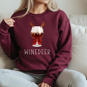 May include: A maroon long-sleeve sweatshirt featuring a graphic of a wine glass with reindeer antlers, a string of small yellow lights, and a red bow. The text WINEDEER is printed in white capital letters below the wine glass illustration.