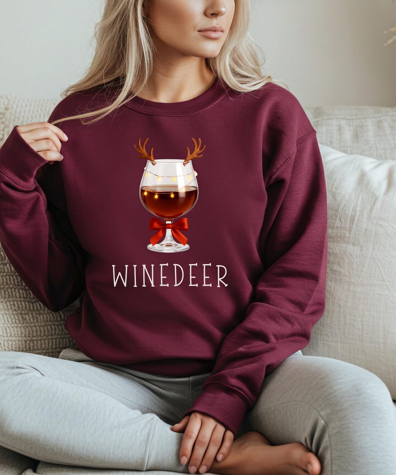May include: A maroon long-sleeve sweatshirt featuring a graphic of a wine glass with reindeer antlers, a string of small yellow lights, and a red bow. The text WINEDEER is printed in white capital letters below the wine glass illustration.