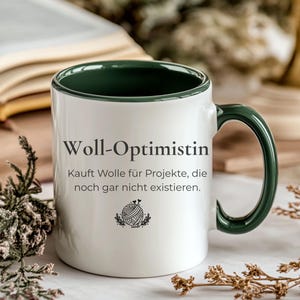Può includere: Una tazza in ceramica bianca con interno e manico verde scuro. La parte anteriore presenta il testo nero "Woll-Optimistin" seguito da "Kauft Wolle für Projekte, die noch gar nicht existieren" e una piccola grafica nera di un gomitolo di lana con ferri da maglia.