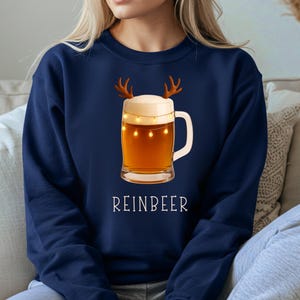 May include: A navy blue crewneck sweatshirt featuring a graphic of a glass beer mug with a thick white foam head, brown reindeer antlers, and a string of small yellow fairy lights wrapped around the glass, with the text REINBEER printed below.
