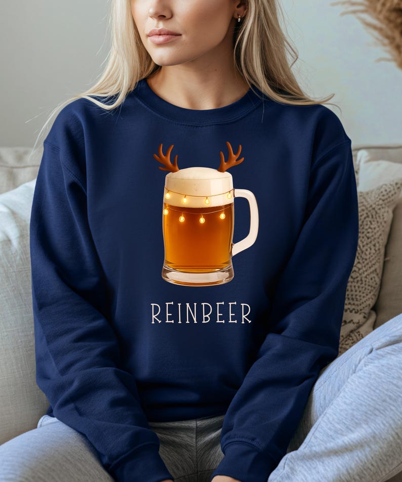 May include: A navy blue crewneck sweatshirt featuring a graphic of a glass beer mug with a thick white foam head, brown reindeer antlers, and a string of small yellow fairy lights wrapped around the glass, with the text REINBEER printed below.