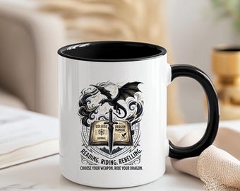 May include: A white ceramic mug with a black handle and interior rim. The front features a graphic of an open book, a sword, and a dragon. Text reads: COLLEGE DRAGON MANUAL, READING. RIDING. REBELLING. CHOOSE YOUR WEAPON, RIDE YOUR DRAGON.