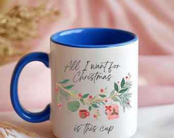May include: A white ceramic mug with a vibrant blue interior and handle. The exterior features the black text All I want for Christmas is this cup, accompanied by a festive illustration of pine branches, red berries, a yarn ball, and a gift.