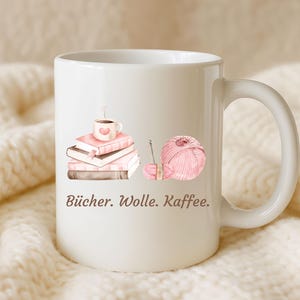 May include: A white ceramic mug featuring a printed illustration of a stack of books topped with a coffee cup, alongside a ball of pink yarn with a knitting needle. The text "B&uuml;cher. Wolle. Kaffee." is printed in brown below the illustrations.
