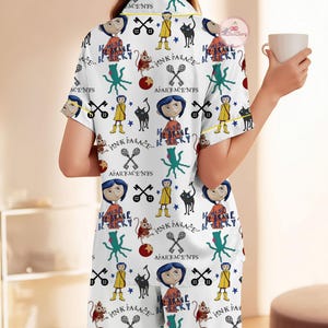 May include: White pyjama set with a repeating pattern of cartoon characters, keys, and text. The short-sleeved top and trousers have yellow trim. The text includes "Pink Palace" and "Be Brave Be Tricky".