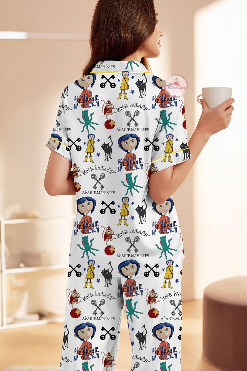 May include: White pyjama set with a repeating pattern of cartoon characters, keys, and text. The short-sleeved top and trousers have yellow trim. The text includes "Pink Palace" and "Be Brave Be Tricky".