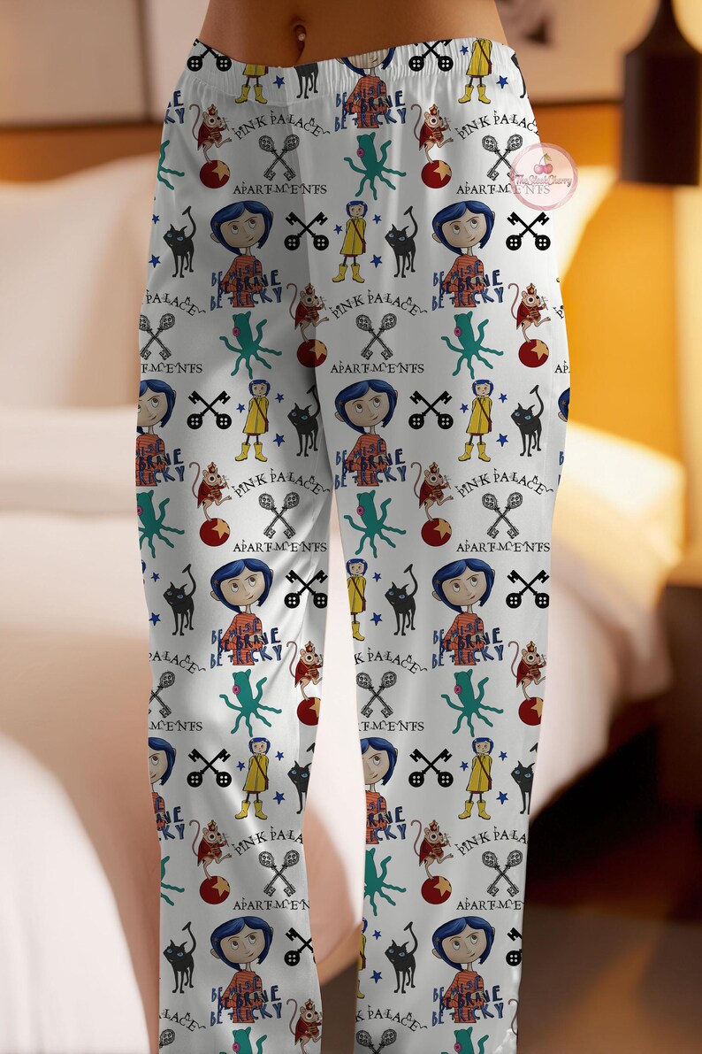 May include: White pajama trousers featuring a repeating pattern of cartoon characters, text, and symbols. The design includes a girl with blue hair, a black cat, and the words "Pink Palace" and "Be Brave".