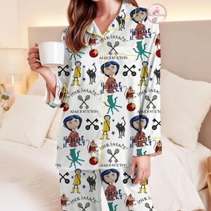 May include: White pyjama set with a repeating pattern of cartoon characters, cats, keys, and the text "PINK PALACE" and "BE BRAVE BE TRICKY". The pyjamas have yellow trim and are worn by a person holding a white mug.
